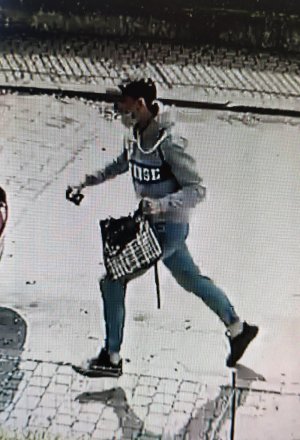 Na zdjęciu widać jak mężczyzna ubrany w siwą sportową bluzę z ciemnym napisem z przodu, niebieskie jeansy ściągane gumką przy kostkach, białe skarpetki, ciemne buty i ciemną czapkę z daszkiem typu bejsbolówka, biegnie z torebką, którą siła wyrwał kobiecie.