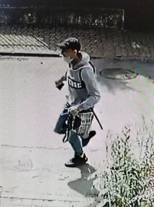 Na zdjęciu widać jak mężczyzna ubrany w siwą sportową bluzę z ciemnym napisem z przodu, niebieskie jeansy ściągane gumką przy kostkach, białe skarpetki, ciemne buty i ciemną czapkę z daszkiem typu bejsbolówka, biegnie z torebką, którą siła wyrwał kobiecie.