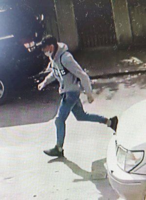 Na zdjęciu widać jak mężczyzna ubrany w siwą sportową bluzę z ciemnym napisem z przodu, niebieskie jeansy ściągane gumką przy kostkach, białe skarpetki, ciemne buty i ciemną czapkę z daszkiem typu bejsbolówka, biegnie z torebką, którą siła wyrwał kobiecie.