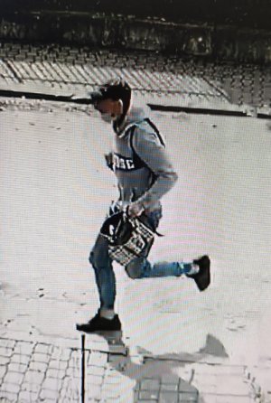 Na zdjęciu widać jak mężczyzna ubrany w siwą sportową bluzę z ciemnym napisem z przodu, niebieskie jeansy ściągane gumką przy kostkach, białe skarpetki, ciemne buty i ciemną czapkę z daszkiem typu bejsbolówka, biegnie z torebką, którą siła wyrwał kobiecie.