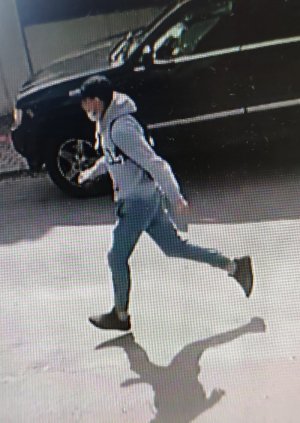 Na zdjęciu widać jak mężczyzna ubrany w siwą sportową bluzę z ciemnym napisem z przodu, niebieskie jeansy ściągane gumką przy kostkach, białe skarpetki, ciemne buty i ciemną czapkę z daszkiem typu bejsbolówka, biegnie z torebką, którą siła wyrwał kobiecie.