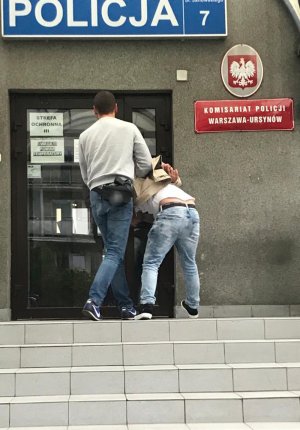 Na zdjęciu, policjant w umundurowaniu cywilnym, doprowadza do Komisariatu Policji Warszawa Ursynów jednego z mężczyzn podejrzanego o kradzież samochodu.  Mężczyzna ma kajdanki założone z tyłu.
