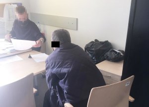 Przy biurku siedzi policjant, który przesłuchuje siedzącego na przeciwko podejrzanego. Funkcjonariusz przekazuje mężczyźnie protokół do zapoznania się z treścią swoich wyjaśnień.