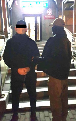 Na zdjęciu wsiadać zatrzymanego mężczyznę ubranego na czarno. Ma założone kajdanki na ręce z przodu. Obok niego stoi policjant, który doprowadza go do Komisariatu Policji.