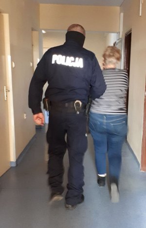 Zdjęcie przedstawia podejrzaną, prowadzoną korytarzem w Komisariacie Policji przez umundurowanego policjanta. Kobieta ma kajdanki założone na ręce z przodu.