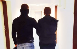 Zdjęcie przedstawia policjanta umundurowanego po cywilnemu, który prowadzi korytarzem za trzymanego 22-latka. Podejrzany ma kajdanki założone na ręce z przodu. Policjant i zatrzymany są odwróceni tyłem do obiektywu.