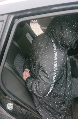 Zdjęcie przedstawia mężczyznę ubranego w czarna kurtce z kapturem siedzącego na tylnym siedzeniu nieoznakowanego, policyjnego radiowozu. Zatrzymany ma na rękach kajdanki założone z tyłu.