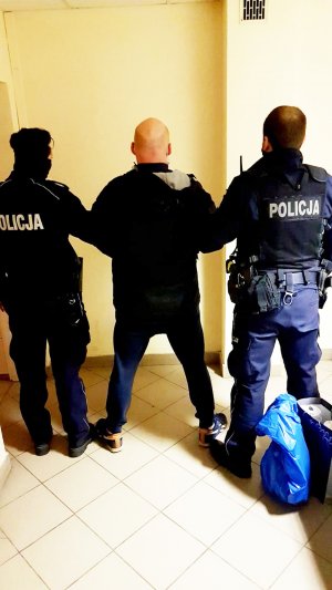 Zdjęcia przedstawia zatrzymanego mężczyznę ubranego w ciemną odzież. Mężczyzna jest odwrócony tyłem do obiektywu. Na rękach ma kajdanki założone z przodu. Obok niego stoją umundurowani funkcjonariusze Policji, którzy go zatrzymywali i doprowadzili do jednostki.