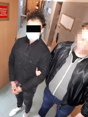 Zdjęcie przedstawia podejrzanego mężczyznę ubranego na czarno, z kajdankami założonymi na ręce z przodu w towarzystwie policjanta po cywilnemu.