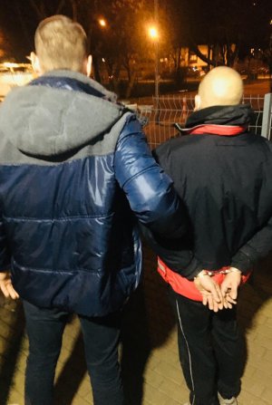 Na zdjęciu widać policjanta umundurowanego po cywilnemu, który doprowadza podejrzanego mężczyznę. Podejrzany ma założone kajdanki na ręce z tyłu.