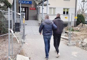 Zdjęcie przedstawia policjanta umundurowanego po cywilnemu, doprowadzającego zatrzymanego mężczyznę do komisariatu Policji. Podejrzany ma kajdanki założone na ręce z przodu.