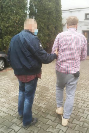 Zdjęcie przedstawia policjanta-operacyjnego, który prowadzi podejrzanego ubranego w kraciastą koszulę i jeansy. Zatrzymany mężczyzna ma założone kajdanki na ręce z przodu.