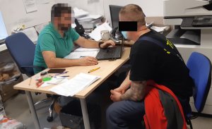 Zdjęcie przedstawia policjanta siedzącego za biurkiem i przesłuchującego podejrzanego. Na przeciw niego na krześle siedzi mężczyzna, który jest przesłuchiwany w charakterze podejrzanego.