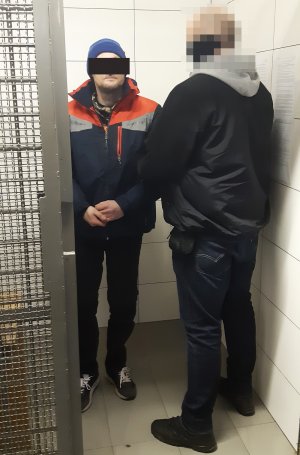 Zdjęcie przedstawia policjanta z zatrzymanym mężczyzną, który ma założone kajdanki na ręce trzymane z przodu.