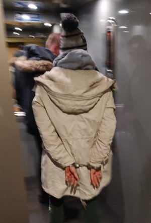 Zdjęcie przedstawia policjanta umundurowanego po cywilnemu doprowadzającego zatrzymana kobietę. Podejrzana ma kajdanki założone na ręce z tyłu.
