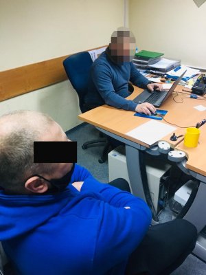 Zdjęcie przedstawia policjanta, siedzącego za biurkiem który wykonuje czynności z siedzącym obok zatrzymanym. podejrzany ma założone kajdanki na ręce z przodu.
