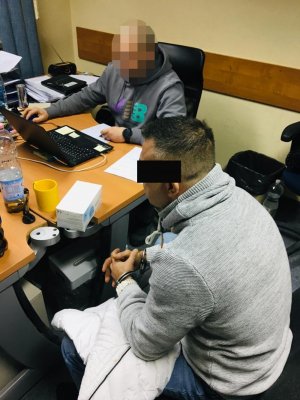 Zdjęcie przedstawia policjanta, siedzącego za biurkiem który wykonuje czynności z siedzącym obok zatrzymanym. podejrzany ma założone kajdanki na ręce z przodu.