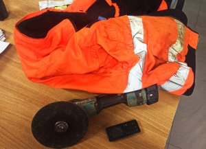 Zdjęcie przedstawia pomarańczową kurtkę z odblaskami, szlifierkę kątowa oraz telefon komórkowy.