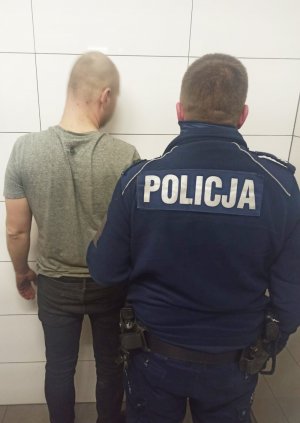 Zdjęcie przedstawia umundurowanego policjanta oraz stojącego obok zatrzymanego mężczyznę.