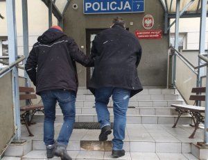 Zdjęcie przedstawia nieumundurowanego policjanta prowadzącego pierwszego z zatrzymanych mężczyzn. 51-latek ma kajdanki założone z przodu.