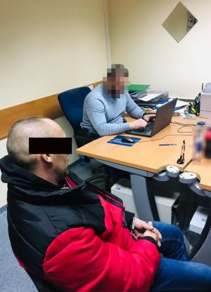 Zdjęcie przedstawia zatrzymanego 42-latka z kajdankami założonymi na ręce z przodu, siedzącego na krześle obok biurka policjanta z wydziału kryminalnego, który wykonuje z nim czynności.