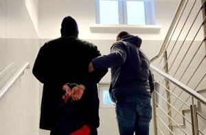 Zdjęcie przedstawia zatrzymanego mężczyznę z kajdankami załojonymi na ręce z tyłu, prowadzonego przez nieumundurowanego policjanta po schodach.