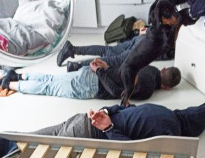 Zdjęcie przedstawia trzech zatrzymanych obywateli Ukrainy podejrzanych o szereg przestępstw, w tym narkotykowych. Trzej mężczyźni leżą na podłoże w szortach oraz t-shirtach. Mają założone kajdanki na ręce z tylu. Nad nimi stoi umundurowany funkcjonariusz Policji.