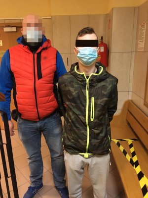 Na zdjęciu nieumundurowany policjant doprowadza podejrzanego mężczyznę na rozprawę sądową. Obaj stoją na korytarzy przed rozprawą. Podejrzany mężczyzna ma kajdanki założone na ręce z tyłu.