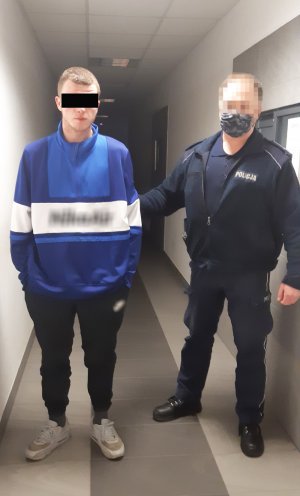 Z prawe strony umundurowany policjant. Z lewej zatrzymany mężczyzna. Funkcjonariusz prowadzi go korytarzem w Komendzie Policji.