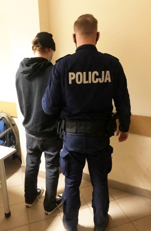 Po prawej stronie stoi umundurowany funkcjonariusz Policji. Obok niego, z jego lewe strony stoi zatrzymany mężczyzna. Podejrzany ma kajdanki założone na ręce z przodu.