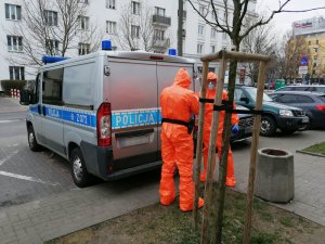 Na zdjęciu policjanci wyposażeni w kombinezony, maseczki oraz rękawiczki sanitarne doprowadzają do budynku komendy Policji przy ul. Malczewskiego w Warszawie zatrzymanego mężczyznę. Podejrzany ma założone kajdanki na ręce z przodu.