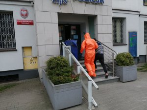 Na zdjęciu policjanci wyposażeni w kombinezony, maseczki oraz rękawiczki sanitarne doprowadzają do budynku komendy Policji przy ul. Malczewskiego w Warszawie zatrzymanego mężczyznę. Podejrzany ma założone kajdanki na ręce z przodu.