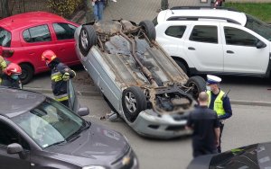 Zdjęcie przedstawia samochód marki mazda wywrócony do góry kołami oraz policjanta ze stołecznej drogówki zabezpieczającego miejsca kolizji drogowej.