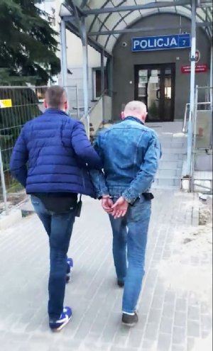 Po lewej stronie nieumundurowany policjant prowadzi podejrzanego mężczyznę do Komisariatu Policji Warszawa Ursynów. Mężczyzna ma kajdanki założone na ręce z tyłu.