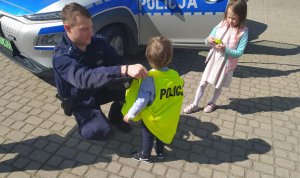 Umundurowani policjanci rozmawiają z przedszkolakami na temat bezpieczeństwa. dzieci przymierzają czapkę, kamizelkę odblaskową, zapoznają się z wyposażeniem dzielnicowych do służby oraz wsiadają do policyjnego radiowozu.