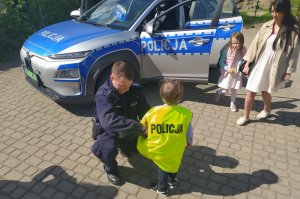 Umundurowani policjanci rozmawiają z przedszkolakami na temat bezpieczeństwa. dzieci przymierzają czapkę, kamizelkę odblaskową, zapoznają się z wyposażeniem dzielnicowych do służby oraz wsiadają do policyjnego radiowozu.