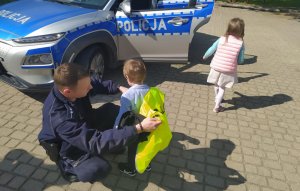 Umundurowani policjanci rozmawiają z przedszkolakami na temat bezpieczeństwa. dzieci przymierzają czapkę, kamizelkę odblaskową, zapoznają się z wyposażeniem dzielnicowych do służby oraz wsiadają do policyjnego radiowozu.