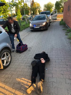 Na zdjęciach widać nieumundurowanych policjantów w trakcie zatrzymania dwóch mężczyzn poruszających się samochodem ford focus. Jeden z zatrzymanych mężczyzn siedzi w pobliżu auta którym kierował. Ma kajdanki założone na rękach z tyłu. Drugi leży na brzuchu i ma kajdanki założone na ręce z tyłu. Policjanci przeszukają ich samochód w poszukiwaniu dowodów na ich przestępcza działalność.