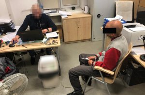 Po lewej stronie, za biurkiem nieumundurowany policjant, który wykonuje czynności z podejrzanym mężczyzną. Podejrzany po prawej stronie. Siedzi na krześle ma kajdanki na rękach z przodu.