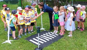 Policjantka rozmawia z dziećmi, pokazując im znaki drogowe. Tłumaczy najmłodszym na czym polega bezpieczeństwo i jakie zasady należy spełniać, aby dzieci były bezpieczne na drodze, w domu, w sieci i podczas kontaktu z agresywnym psem.