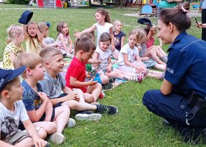 Policjantka rozmawia z dziećmi, pokazując im znaki drogowe. Tłumaczy najmłodszym na czym polega bezpieczeństwo i jakie zasady należy spełniać, aby dzieci były bezpieczne na drodze, w domu, w sieci i podczas kontaktu z agresywnym psem.