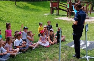 Policjantka rozmawia z dziećmi, pokazując im znaki drogowe. Tłumaczy najmłodszym na czym polega bezpieczeństwo i jakie zasady należy spełniać, aby dzieci były bezpieczne na drodze, w domu, w sieci i podczas kontaktu z agresywnym psem.