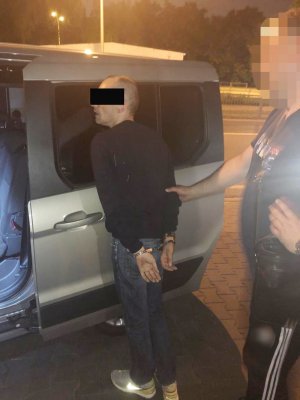 Na zdjęciu nieumundurowany policjant z zatrzymanym, który ma kajdanki założone na ręce z tyłu.