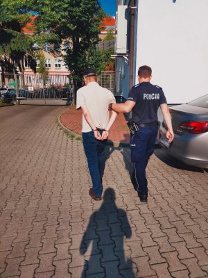 Po prawej stronie umundurowany policjant prowadzi znajdującego się po lewej stronie zatrzymanego mężczyznę. Poszukiwany ma kajdanki założone na ręce z tyłu.