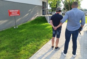 Po prawej stronie nieumundurowany policjant. Po lewej stronie podejrzany mężczyzna prowadzony przez policjanta do Komisariatu Policji Warszawa Ursynów. 52-latek ma kajdanki założone na ręce z tyłu.