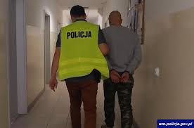 Policjant z zatrzymanym mężczyzną, prowadzi go korytarzem w budynku. Zatrzymany ma założone kajdanki.