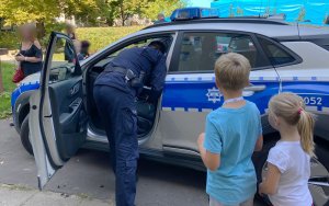 Policjant umundurowany rozmawiający z uczestnikami pikniku stoi obok radiowozu policyjnego.
