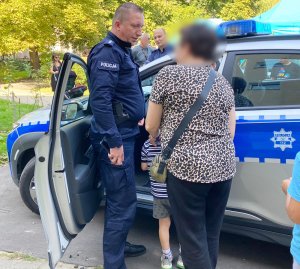 Policjant umundurowany rozmawiający z uczestnikami pikniku stoi obok radiowozu policyjnego.