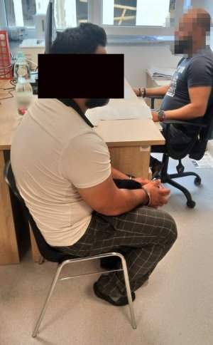 Policjant wykonuje czynności z mężczyzną siedzącym przy biurku.