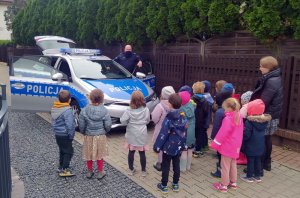 Na zdjęciu dzielnicowy prowadzi zajęcia z dziećmi z Przedszkola Frajda na warszawskim Wilanowie  mówiąc o tym, jak postępować, aby dni mijały bezpiecznie w domu, w przedszkolu, w świecie wirtualnym i na drodze.